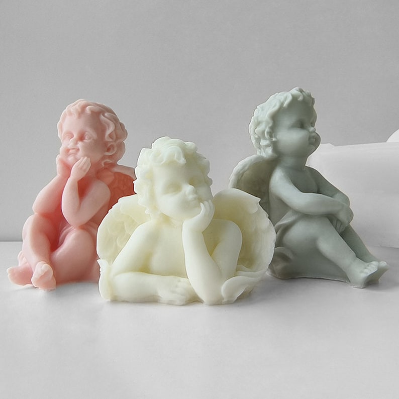 May include: Three cherub figurines made of white, pink and grey resin. The cherubs are sitting with their wings spread out.