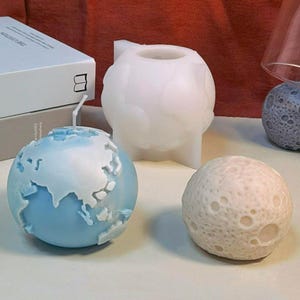Puede incluir: Tres velas redondas con forma de planetas. La primera vela es un globo terráqueo azul con continentes blancos. La segunda vela es una esfera blanca con una superficie texturizada. La tercera vela es una esfera marrón claro con una superficie texturizada.