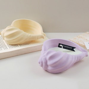 May include: Two seashell-shaped candles, one in a pale yellow colour and the other in a pale purple colour. The purple candle has a label that reads "HANDMADE" with a small wick sticking out of the top.
