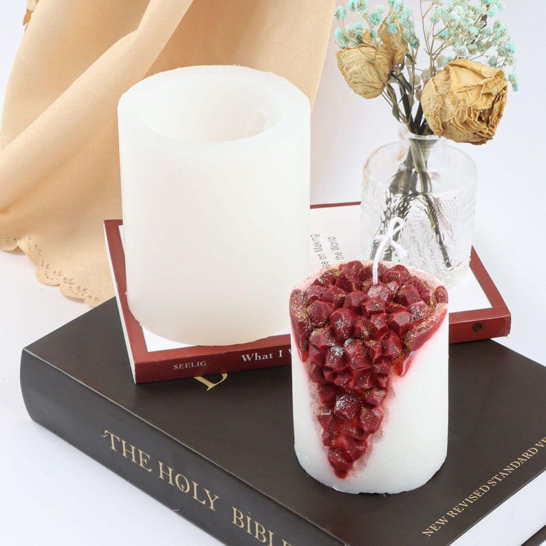 May include: A white candle with a red, glittery, grape-shaped design. The candle is sitting on a book with the words "The Holy Bible" printed on the cover. The candle is in a white silicone mould.