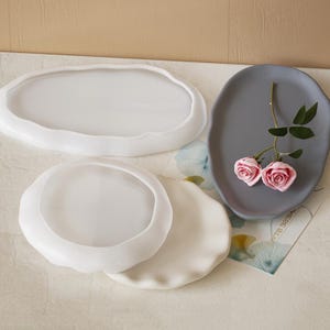 May include: Three white silicone trays, two round and one oval, with a grey silicone tray with a wavy edge. The grey tray has two pink roses on it.