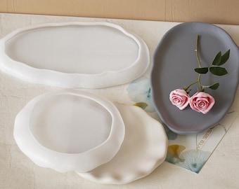 Può includere: Tre vassoi in silicone bianco, due rotondi e uno ovale, con un vassoio in silicone grigio con bordo ondulato. Il vassoio grigio ha due rose rosa sopra.