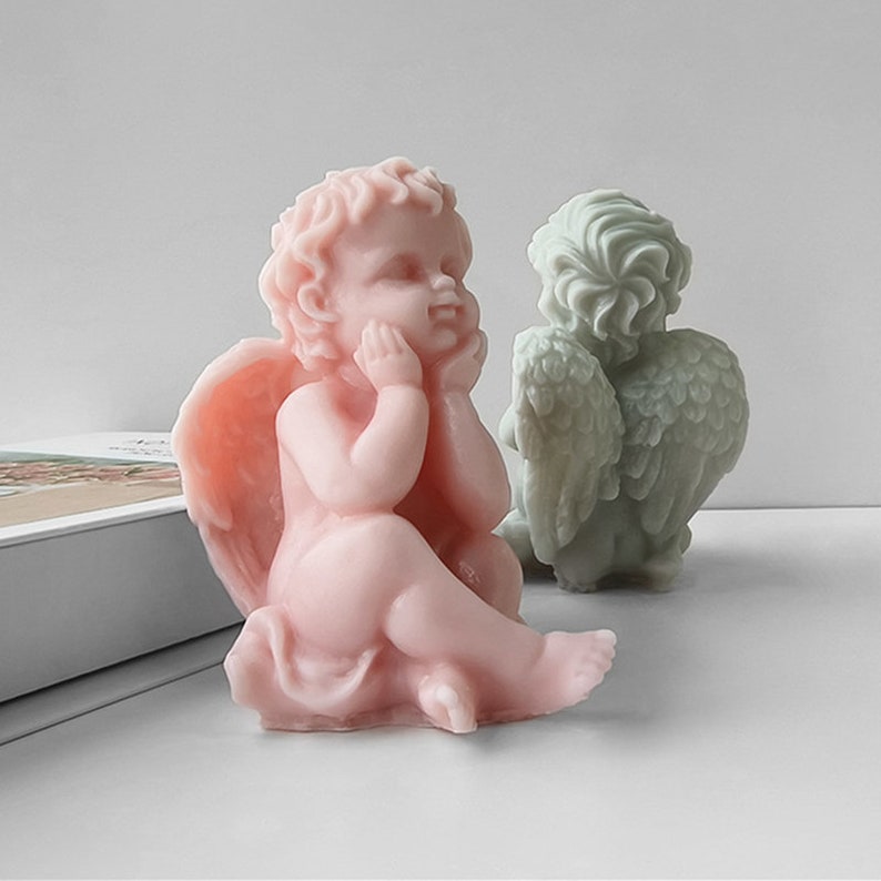 May include: Two cherub figurines, one in pink and one in green, are sitting with their wings spread out. The pink cherub is sitting with its head resting on its hand, while the green cherub is sitting with its wings spread out behind it.