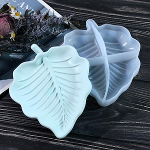 May include: Two light blue silicone leaf-shaped molds. One mold is a single leaf shape, the other is divided into three sections.