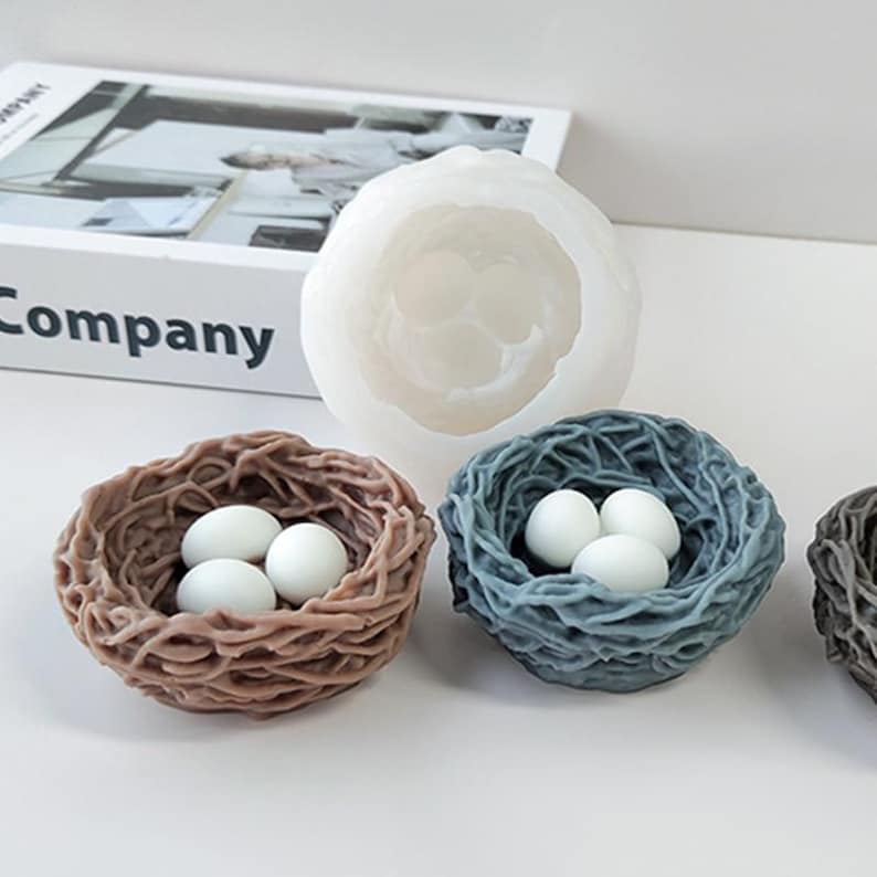 Puede incluir: Tres velas blancas en forma de huevo en un nido de p&aacute;jaro tejido marr&oacute;n, un nido de p&aacute;jaro tejido azul y un molde de silicona blanco de un nido de p&aacute;jaro.