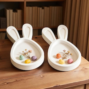 May include: Two white ceramic bunny-shaped dishes with raised ears. Each dish contains colourful Easter eggs and dried flowers. The dishes are on a wooden surface, ideal for Easter decorations.