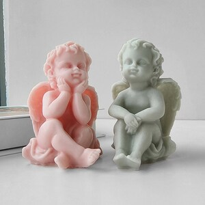 May include: Two small angel figurines, one in pink and one in green, are sitting with their wings spread out behind them. They are both looking down with a thoughtful expression on their faces.