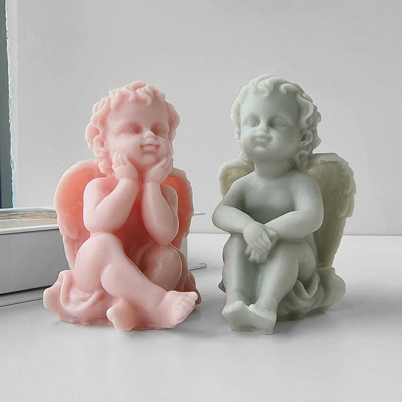 May include: Two small angel figurines, one in pink and one in green, are sitting with their wings spread out behind them. They are both looking down with a thoughtful expression on their faces.