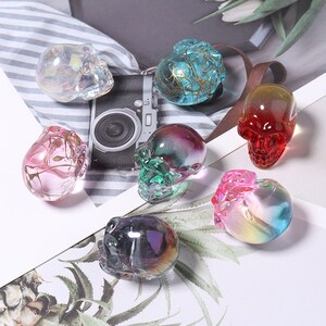 K&ouml;nnte beinhalten: Sieben Kristallsch&auml;del-Figuren in verschiedenen Farben, darunter rosa, blau, gr&uuml;n und schillernd. Die Sch&auml;del sind aus Glas und haben eine glatte, polierte Oberfl&auml;che.