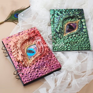 Pu&ograve; includere: Due diari a spirale con copertine iridescenti che presentano un design a occhio di drago. Un diario ha una copertina verde con un occhio dorato e l'altro ha una copertina rosa con un occhio blu.