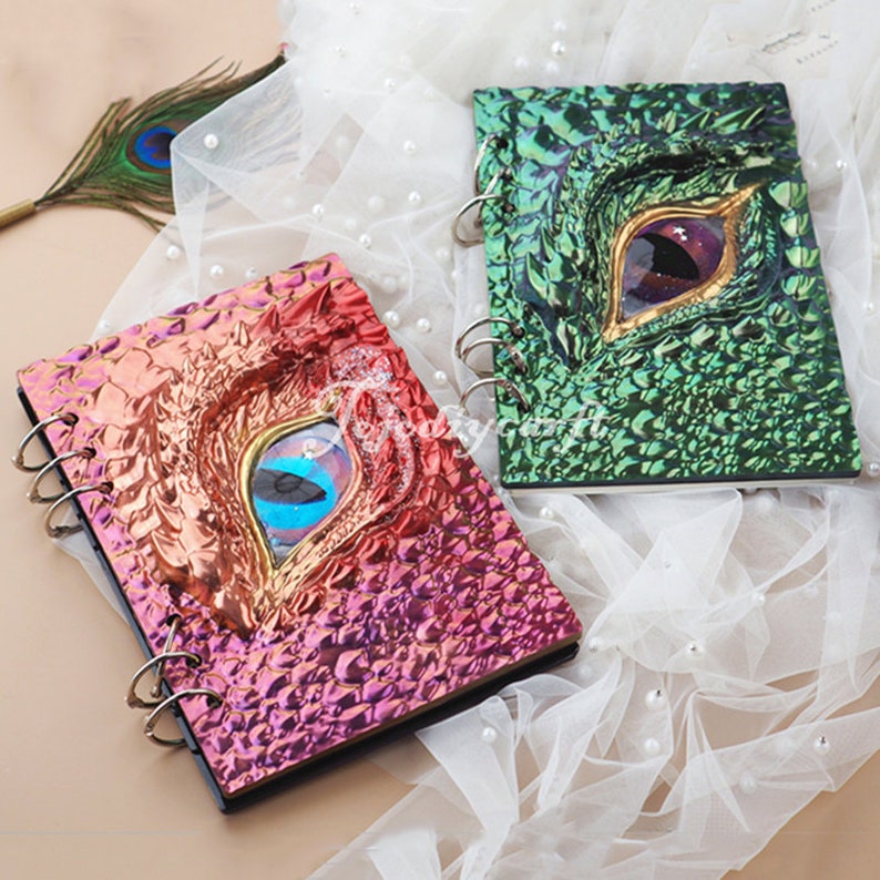 Pu&ograve; includere: Due diari a spirale con copertine iridescenti che presentano un design a occhio di drago. Un diario ha una copertina verde con un occhio dorato e l'altro ha una copertina rosa con un occhio blu.
