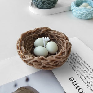 Puede incluir: Una vela de nido de p&aacute;jaro marr&oacute;n con tres huevos blancos en su interior. El nido est&aacute; hecho de cera tejida y los huevos tambi&eacute;n son de cera.