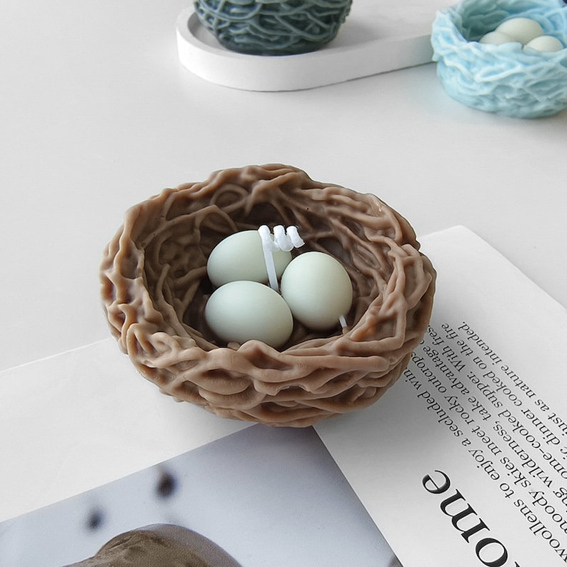 Puede incluir: Una vela de nido de p&aacute;jaro marr&oacute;n con tres huevos blancos en su interior. El nido est&aacute; hecho de cera tejida y los huevos tambi&eacute;n son de cera.