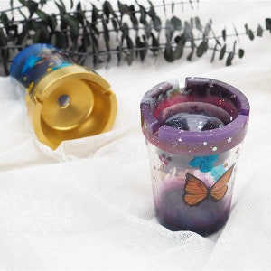 May include: Two decorative ashtrays with a galaxy design. One ashtray is gold with a blue and white galaxy design. The other ashtray is purple with a galaxy design and a butterfly.