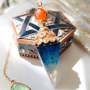 May include: A blue and white crystal pendulum with a gold-toned top and an orange gemstone. The pendulum is hanging from a gold chain.