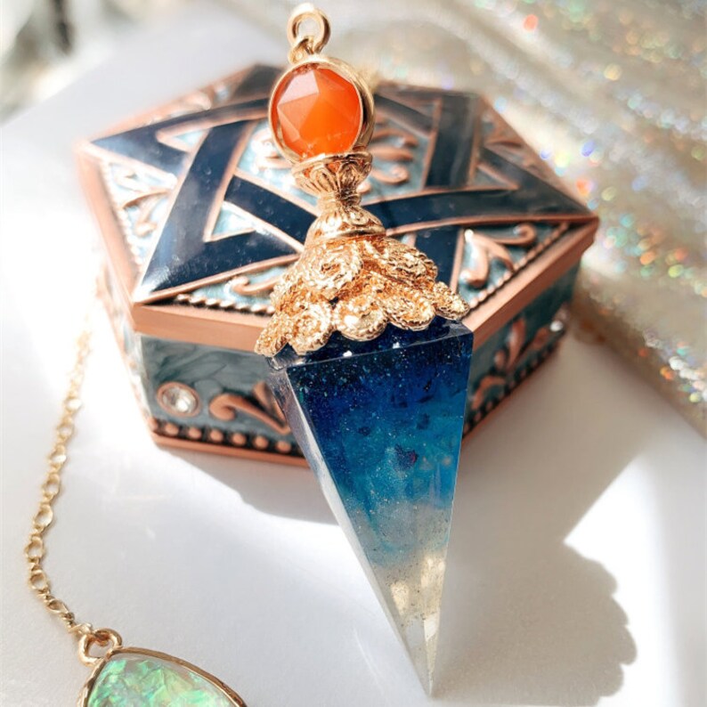 May include: A blue and white crystal pendulum with a gold-toned top and an orange gemstone. The pendulum is hanging from a gold chain.