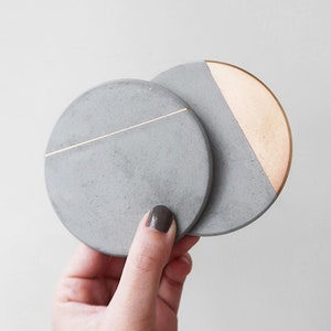 May include: Two round concrete coasters with a gold accent. One coaster has a thin gold line across the top, and the other has a gold half-circle on the side.