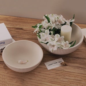 Könnte beinhalten: Zwei cremefarbene, gerippte Keramikschalen auf einer Holzoberfläche. Eine Schale enthält weiße Blumen und eine Kerze. Die andere Schale ist leer, mit einem einzelnen Blütenblatt darin. Eine Karte mit "JUST FOR YOU" ist ebenfalls vorhanden.