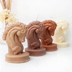 May include: Four decorative horse head candles in different shades of brown and white. The candles are sculpted in a realistic style and are arranged in a row on a white surface.