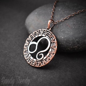 May include: A copper and silver wire wrapped pendant necklace featuring the Leo zodiac sign. The pendant is round and has a woven design.