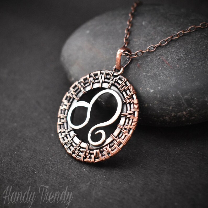 May include: A copper and silver wire wrapped pendant necklace featuring the Leo zodiac sign. The pendant is round and has a woven design.
