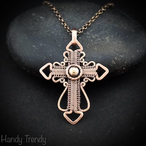 May include: Copper wire-wrapped cross pendant necklace. The handcrafted cross features a central silver-colored bead and intricate wire detailing. The pendant hangs from a delicate copper chain. The necklace is displayed on a dark stone.