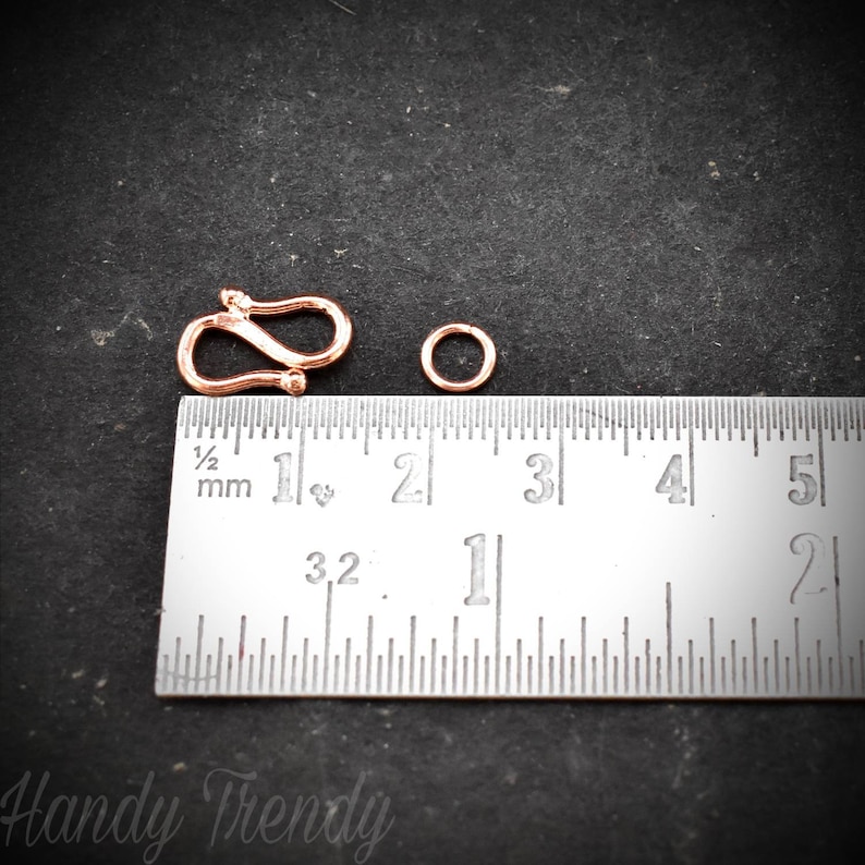 May include: A close-up of a gold-toned metal clasp and jump ring. The clasp is shaped like an 'S' and measures approximately 2.5 cm in length. The jump ring is about 1.25 cm in diameter. A ruler is visible in the background.