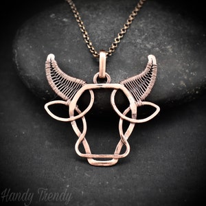 May include: A copper wire bull pendant necklace with a Celtic knot design. The pendant is hanging from a chain on a dark grey background.