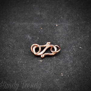 May include: A rose gold-coloured metal clasp for jewellery making. The clasp is shaped like a figure eight and has a smooth, polished finish.