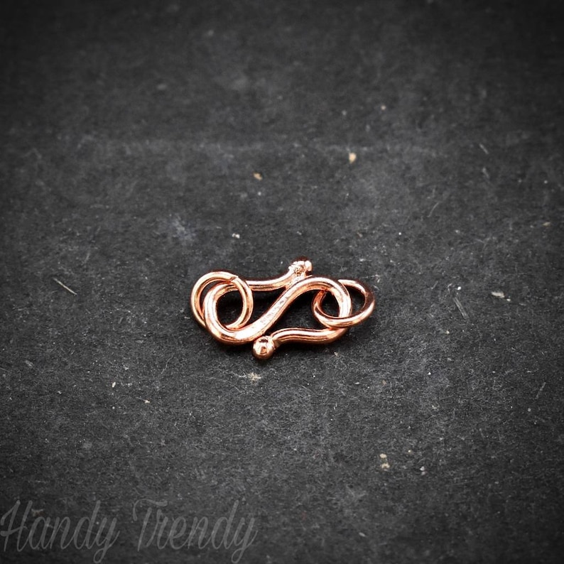 May include: A rose gold-coloured metal clasp for jewellery making. The clasp is shaped like a figure eight and has a smooth, polished finish.