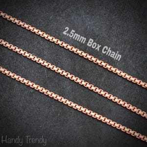 Può includere: Primo piano di tre catene a scatola color rame da 2,5 mm. Le catene sono disposte diagonalmente su uno sfondo scuro. Il testo "2.5mm Box Chain" è visualizzato in bianco. È visibile anche il marchio "Handy Trendy".