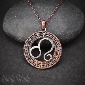 May include: A copper chain necklace with a silver Leo zodiac sign pendant. The pendant is surrounded by a woven copper wire design.