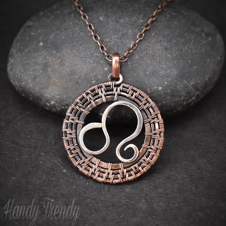 May include: A copper chain necklace with a silver Leo zodiac sign pendant. The pendant is surrounded by a woven copper wire design.