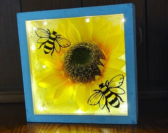 May include: A light blue wooden frame with a clear glass front, containing a yellow sunflower with two black bees. The frame is illuminated with warm white LED lights.