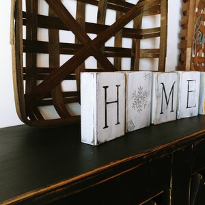 May include: White wooden blocks spelling "HOME" with a snowflake on the "O" block and a painted pumpkin on the "E" block. The blocks are sitting on a black surface.