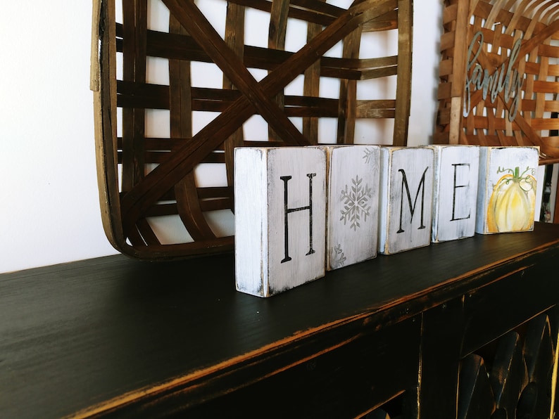 May include: White wooden blocks spelling "HOME" with a snowflake on the "O" block and a painted pumpkin on the "E" block. The blocks are sitting on a black surface.