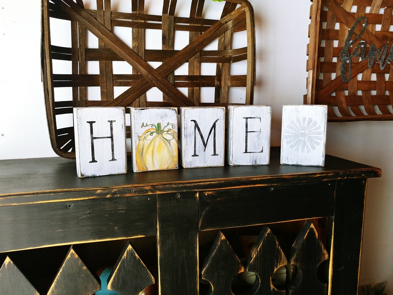 May include: White wooden blocks spelling "HOME" with a painted pumpkin on the "O" block. The blocks are sitting on a black distressed wood surface.