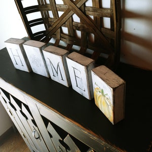 May include: White wooden blocks spelling "HOME" with a painted pumpkin on the last block. The blocks are sitting on a black painted table with a distressed finish.