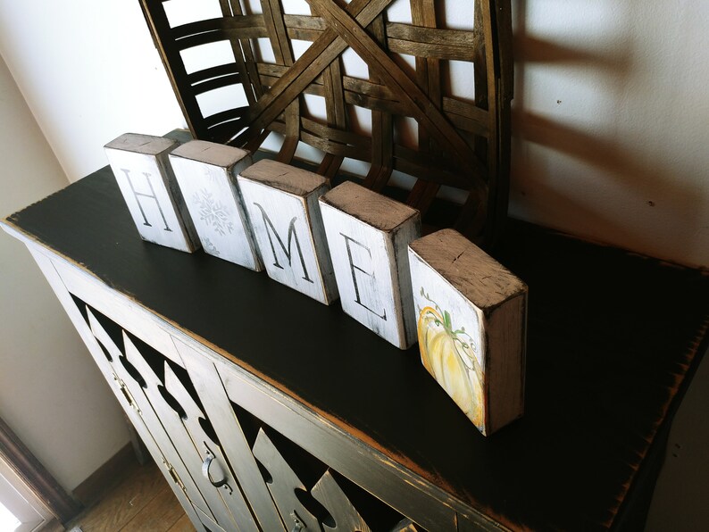 May include: White wooden blocks spelling "HOME" with a painted pumpkin on the last block. The blocks are sitting on a black painted table with a distressed finish.