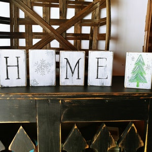 May include: Wooden blocks spelling "HOME" with a snowflake design on one block and a green Christmas tree on another. The blocks are sitting on a black distressed table.