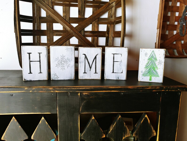 May include: Wooden blocks spelling "HOME" with a snowflake design on one block and a green Christmas tree on another. The blocks are sitting on a black distressed table.