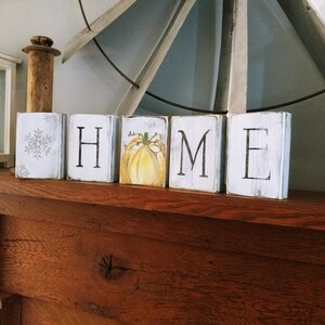 May include: Wooden blocks spelling out the word "HOME" with a painted pumpkin on the "O" block. The blocks are white with black lettering and are sitting on a wooden surface.