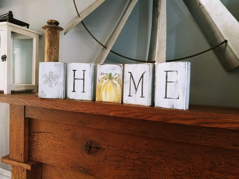 May include: Wooden blocks spelling out the word "HOME" with a painted pumpkin on the "O" block. The blocks are white with black lettering and are sitting on a wooden surface.