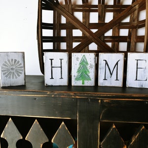 May include: Wooden blocks spelling "HOME" with a painted green Christmas tree on the "O" block. The blocks are sitting on a black wooden surface.