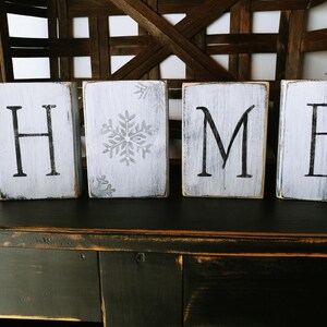May include: White wooden blocks spelling out the word "HOME" with a snowflake design on the second block. The blocks are sitting on a dark wooden surface.