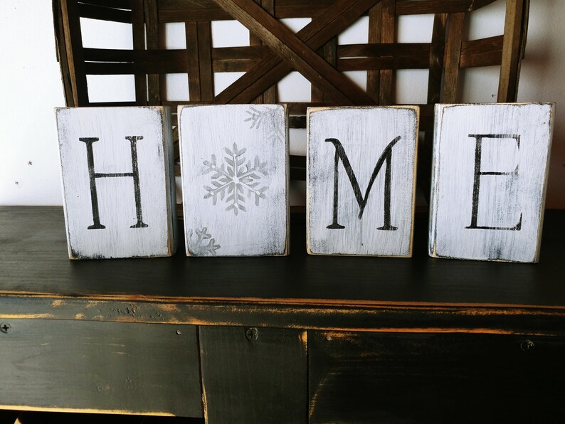 May include: White wooden blocks spelling out the word "HOME" with a snowflake design on the second block. The blocks are sitting on a dark wooden surface.