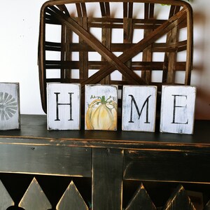 May include: White wooden blocks spelling "HOME" with a painted pumpkin on the "O" block. The blocks are sitting on a black distressed wood surface.