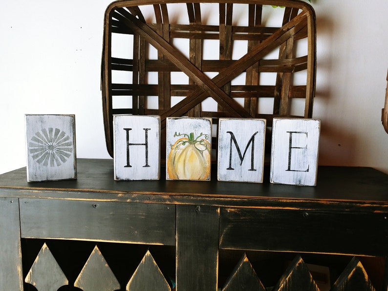 May include: White wooden blocks spelling "HOME" with a painted pumpkin on the "O" block. The blocks are sitting on a black distressed wood surface.
