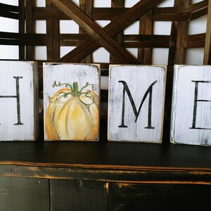 May include: Four wooden blocks spelling out the word "HOME" with a painted pumpkin on the second block. The blocks are white with black lettering and a distressed finish.