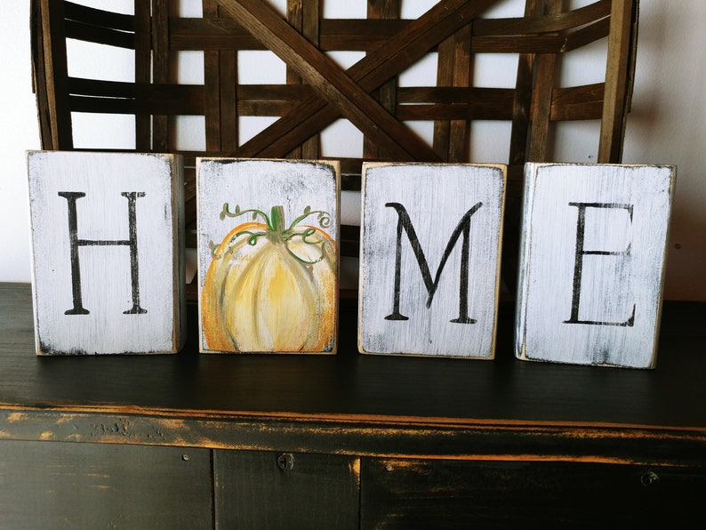 May include: Four wooden blocks spelling out the word "HOME" with a painted pumpkin on the second block. The blocks are white with black lettering and a distressed finish.